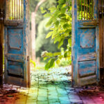 Picture of a rainbow path with two old wood doors opening. The door are painted blue but it's worn and chipped. Beyond the doors is light and beautiful greenery. A photographic depiction of pet loss grief.